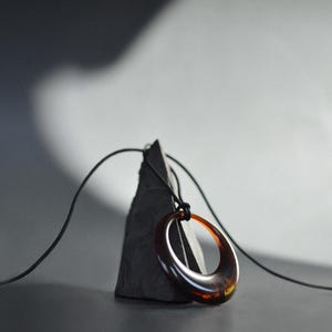 Op de afbeelding: Een minimalistische ketting met een grote, ovale hanger in bruine en amberkleurige tinten. De hanger hangt aan een dun zwart koord. De ketting rust op een donkere, driehoekige steen tegen een neutrale achtergrond.