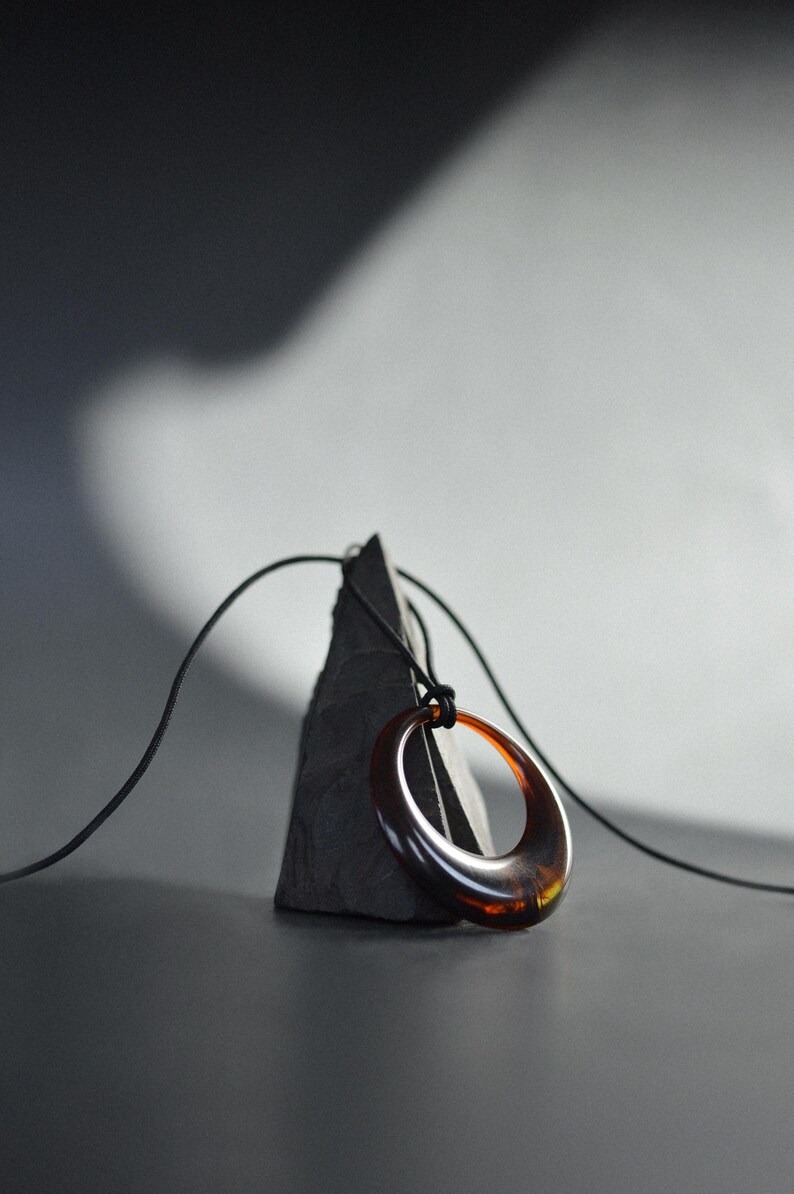 Op de afbeelding: Een minimalistische ketting met een grote, ovale hanger in bruine en amberkleurige tinten. De hanger hangt aan een dun zwart koord. De ketting rust op een donkere, driehoekige steen tegen een neutrale achtergrond.