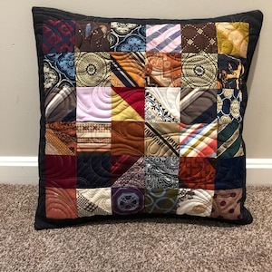 May include: A square throw pillow constructed from a patchwork of various silk neckties in diverse colors and patterns, including geometric, floral, and striped designs. The pillow features a solid black border and intricate swirling machine quilting.