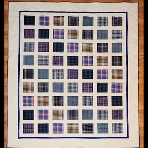 May include: A rectangular quilt featuring a grid of square fabric patches in various plaid patterns, including shades of blue, brown, and red. The patches are separated by cream-colored fabric and framed by a dark blue border and a cream border.