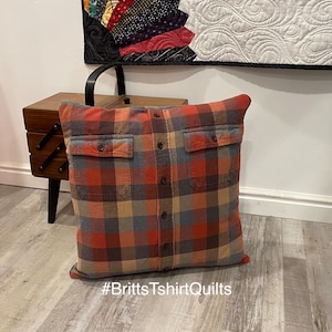 May include: A square throw pillow crafted from a plaid flannel shirt featuring a button-down front placket, two chest pockets with buttoned flaps, and a multi-colored pattern of orange, blue, and tan. The text #BrittsTshirtQuilts is visible.