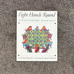 May include: A book titled "Eight Hands Round: A Patchwork Alphabet" features a colorful illustration of children around a patchwork alphabet. The cover art includes the names of the author and illustrator.