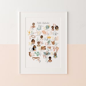 May include: A framed print featuring the "Bible Alphabet" in a colorful, illustrated style. Each letter is paired with a biblical figure or symbol, such as Abraham, Creation, and Jesus. The artwork is set against a two-tone pink and white background.