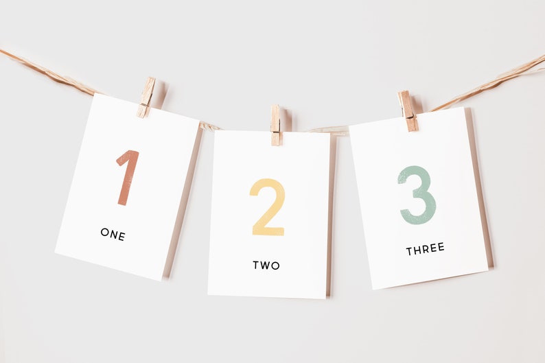 May include: Three white cards with numbers 1, 2, and 3 written in different colors. The cards are hung on a string with clothespins. The numbers are written in a playful, hand-drawn style. The words "one", "two", and "three" are written below the numbers.
