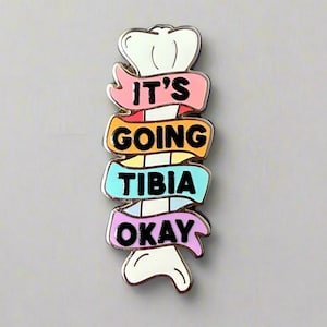 May include: Enamel pin in the shape of a bone with the words "IT'S GOING TIBIA OKAY" on colorful banners. The pin has a silver border and is set against a gray background.