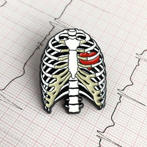May include: A black, white, and tan enamel pin in the shape of a rib cage with a red heart. The pin is set against a pink and white grid background with a black EKG line.