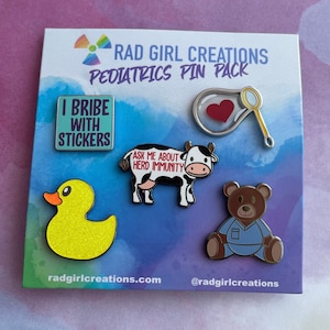 May include: A set of six enamel pins with a paediatric theme. The pins feature a yellow rubber duck, a cow with the text "Ask me about herd immunity", a teddy bear wearing a blue doctor's uniform, a bubble with a red heart inside, a magnifying glass, and a blue and green pin with the text "I bribe with stickers".