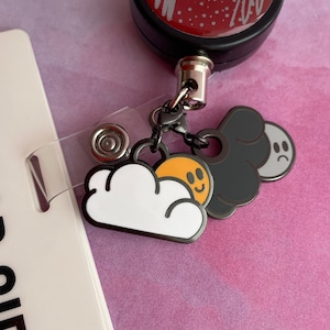 May include: A black retractable badge holder with a silver chain and three enamel charms. The charms are a white cloud with a yellow sun, a black cloud, and a gray moon with a sad face.