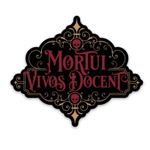 Peut inclure: Un autocollant noir avec l'inscription "Mortui Vivos Docent" en script bordeaux. L'autocollant a une bordure dorée décorative avec des accents de crâne. Le design est orné et de style gothique.
