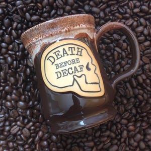 May include: A brown ceramic coffee mug with a handle, featuring the phrase "DEATH BEFORE DECAF" and a skull design. The mug has a two-tone glaze with a lighter brown rim and is set against a background of coffee beans.