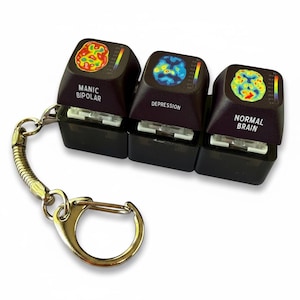 May include: A keychain featuring three black mechanical keyboard keycaps. Each keycap displays a colorful brain scan image and text: Manic Bipolar, Depression, and Normal Brain. The keycaps are attached to a silver metal ring and spring coil.