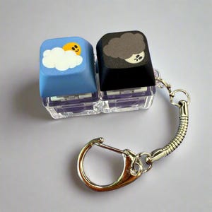 May include: A keychain featuring two keyboard keycaps. One keycap is light blue with a cartoon sun and cloud design, and the other is black with a cloud and moon design. The keychain has a silver clasp and a coiled metal connector.