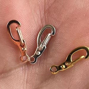 May include: Three small metal clasp hooks in rose gold, silver, and gold tones. The clasps are open, showing the hook shape and the small ring at the base. The clasps are shown against a skin-toned background.