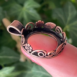 May include: A copper-colored metal crown-shaped pendant with floral and leaf details. The pendant has a loop at the top for hanging and a circular opening in the center. The design features delicate swirls and a dark interior.