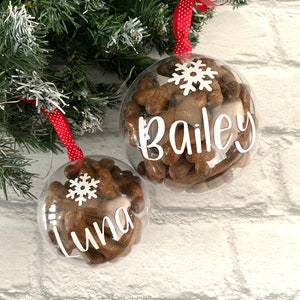 May include: Two clear baubles filled with brown treats, each with a white snowflake and a name in white script: "Bailey" and "Luna." The baubles hang from red ribbons with white polka dots, against a backdrop of green and white Christmas greenery.