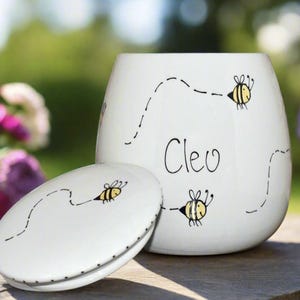 May include: A white ceramic jar with a matching lid, featuring hand-drawn illustrations of yellow and black bees with dotted flight paths. The name Cleo is written in black script on the front of the jar. The container has a smooth, rounded shape.