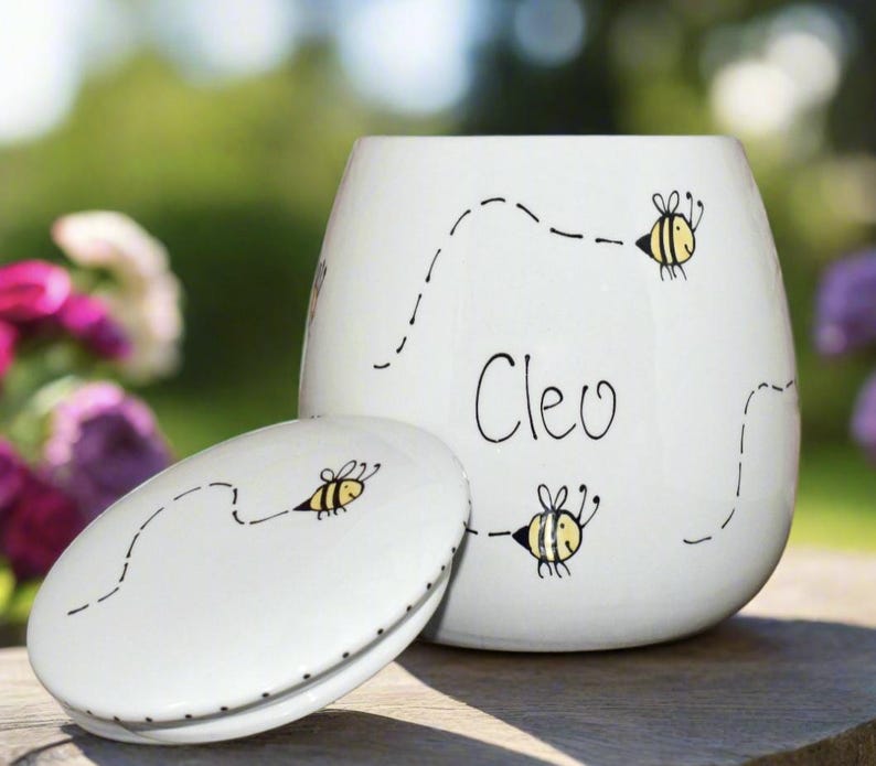 May include: A white ceramic jar with a matching lid, featuring hand-drawn illustrations of yellow and black bees with dotted flight paths. The name Cleo is written in black script on the front of the jar. The container has a smooth, rounded shape.