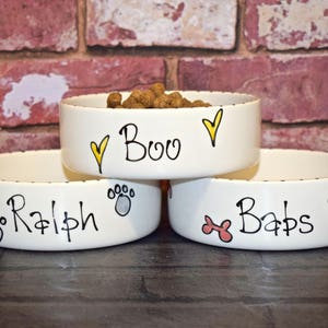 May include: Three white ceramic dog bowls with different names and designs. The top bowl says "Boo" with yellow hearts. The middle bowl says "Ralph" with paw prints. The bottom bowl says "Babs" with bone shapes.
