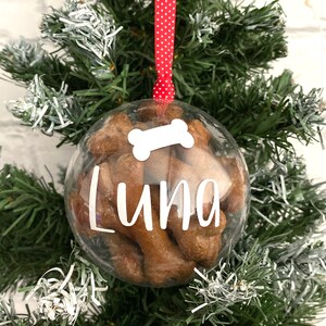 May include: A clear bauble filled with dog treats, suspended by a red and white polka dot ribbon. The bauble features a white bone graphic and the name "Luna" in white script. It is displayed on a green Christmas tree.