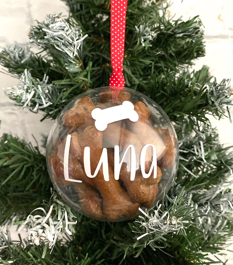 May include: A clear bauble filled with dog treats, suspended by a red and white polka dot ribbon. The bauble features a white bone graphic and the name "Luna" in white script. It is displayed on a green Christmas tree.