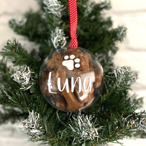 May include: A clear bauble filled with dog treats, featuring a white paw print and the name "Luna" in white script. It hangs from a red and white polka dot ribbon, set against a backdrop of a green Christmas tree, a festive decoration.