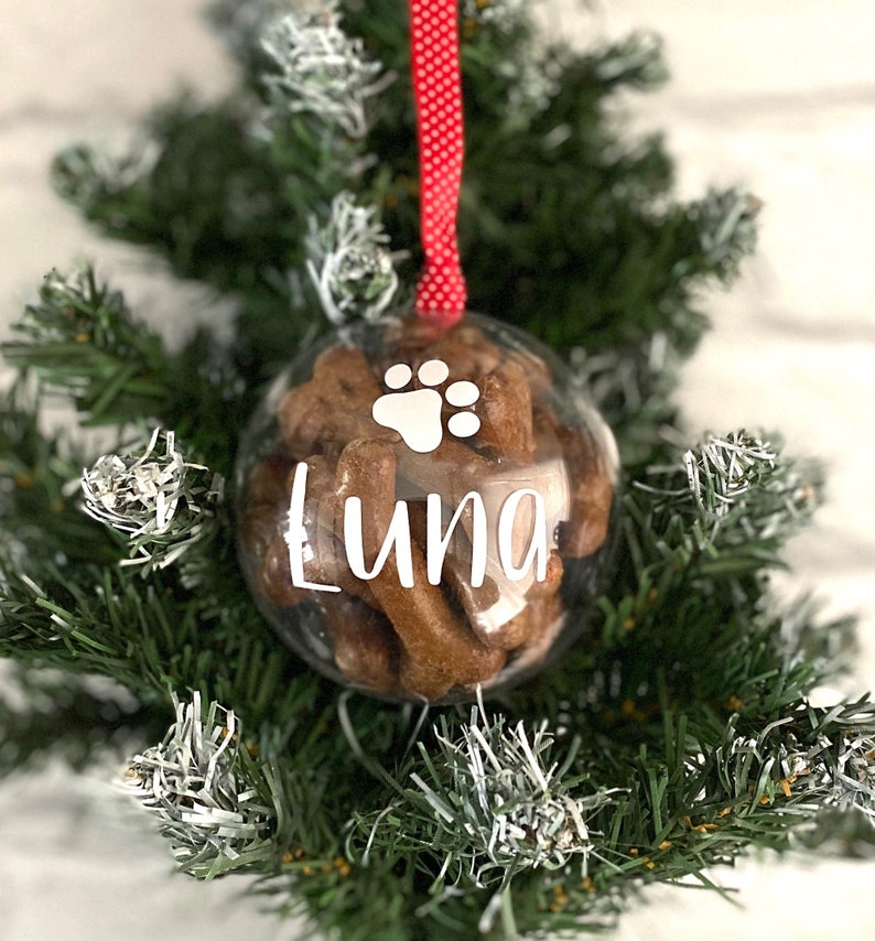 May include: A clear bauble filled with dog treats, featuring a white paw print and the name "Luna" in white script. It hangs from a red and white polka dot ribbon, set against a backdrop of a green Christmas tree, a festive decoration.