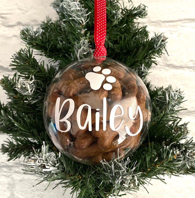 May include: A clear bauble filled with dog treats, with the name "Bailey" in white script. A white paw print is above the name. The bauble hangs from a red ribbon with white polka dots, set against a backdrop of green and white faux evergreen branches.