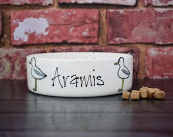May include: White ceramic pet bowl with a black outline and two hand-drawn seagulls. The bowl has the name "Aramis" written in black cursive.