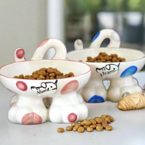 May include: Two ceramic pet bowls with raised paw-shaped bases and curved handles. The front bowl features pink spots and the name Minnie, while the back bowl has blue spots and the name Aramis. Both bowls are filled with brown dry pet food.