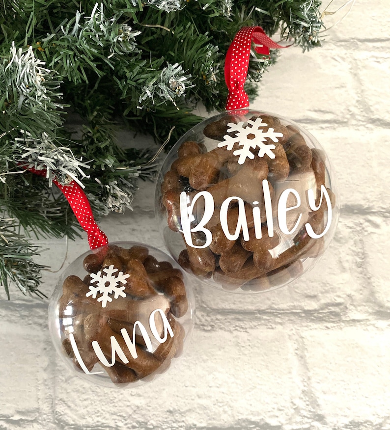 May include: Two clear baubles filled with brown treats, each with a white snowflake and a name in white script: "Bailey" and "Luna." The baubles hang from red ribbons with white polka dots, against a backdrop of green and white Christmas greenery.