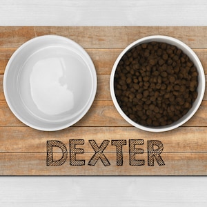 May include: A wooden pet food mat with two white bowls. One bowl is filled with brown dog food. The mat has the name "Dexter" written on it.