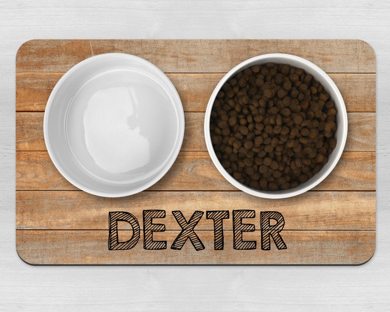 May include: A wooden pet food mat with two white bowls. One bowl is filled with brown dog food. The mat has the name "Dexter" written on it.