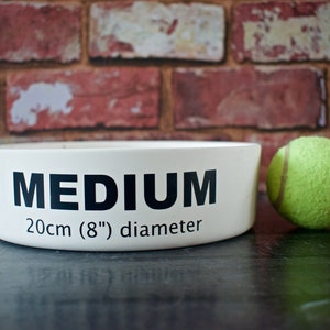 K&ouml;nnte beinhalten: Wei&szlig;e Keramik-Hundenapf mit schwarzem Text "MEDIUM 20cm (8") Durchmesser". Neben der Sch&uuml;ssel liegt ein gr&uuml;ner Tennisball.