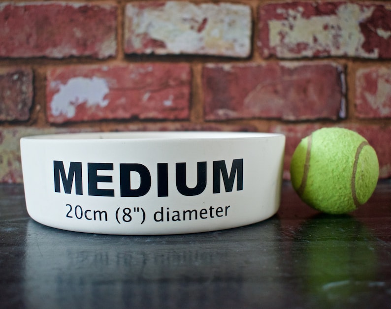 K&ouml;nnte beinhalten: Wei&szlig;e Keramik-Hundenapf mit schwarzem Text "MEDIUM 20cm (8") Durchmesser". Neben der Sch&uuml;ssel liegt ein gr&uuml;ner Tennisball.