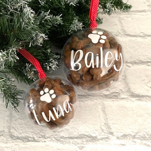 May include: Two clear baubles filled with dog treats, suspended from a Christmas tree. One has the name "Bailey" and a paw print, the other "Luna" and a paw print. They hang by red ribbons with white polka dots.