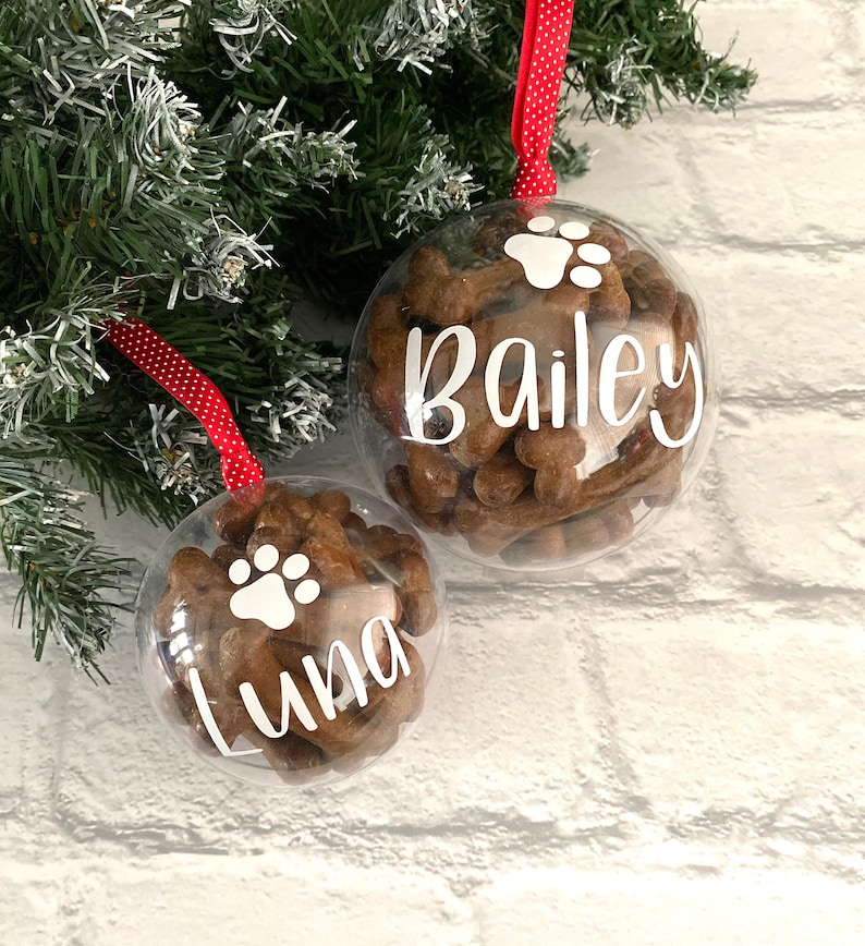 May include: Two clear baubles filled with dog treats, suspended from a Christmas tree. One has the name "Bailey" and a paw print, the other "Luna" and a paw print. They hang by red ribbons with white polka dots.