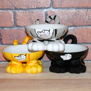 May include: Three ceramic pet bowls shaped like cats with personalized names "Buddy", "Monty", and "Obi". The bowls are in grey, yellow, and black.