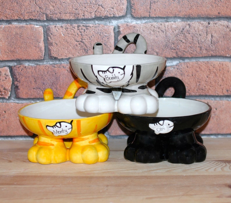 May include: Three ceramic pet bowls shaped like cats with personalized names "Buddy", "Monty", and "Obi". The bowls are in grey, yellow, and black.