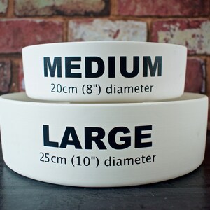 May include: Two white ceramic dog bowls, one stacked on top of the other. The top bowl is labeled "MEDIUM 20cm (8") diameter" and the bottom bowl is labeled "LARGE 25cm (10") diameter".