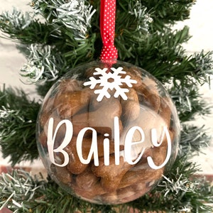 May include: A clear bauble filled with brown dog biscuits, a white snowflake, and the name "Bailey" in white script. The ornament hangs from a red ribbon with white polka dots, set against a green Christmas tree.