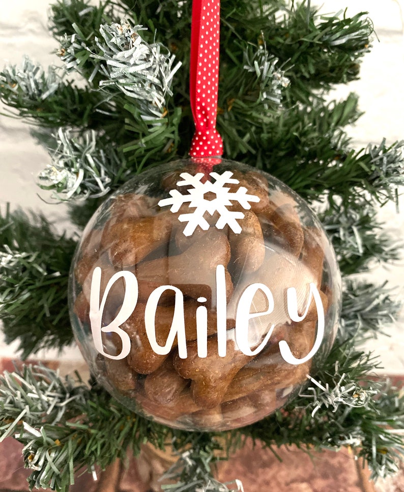 May include: A clear bauble filled with brown dog biscuits, a white snowflake, and the name "Bailey" in white script. The ornament hangs from a red ribbon with white polka dots, set against a green Christmas tree.
