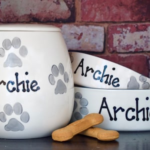 May include: White ceramic pet food and water bowls with the name "Archie" written in black. The bowls are decorated with grey paw prints. There are also dog biscuits on the table.