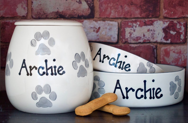May include: White ceramic pet food and water bowls with the name "Archie" written in black. The bowls are decorated with grey paw prints. There are also dog biscuits on the table.