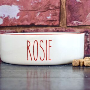 K&ouml;nnte beinhalten: Wei&szlig;e Keramik-Sch&uuml;ssel f&uuml;r Haustiere mit rotem Rand und dem Namen "Rosie" in roten Buchstaben.