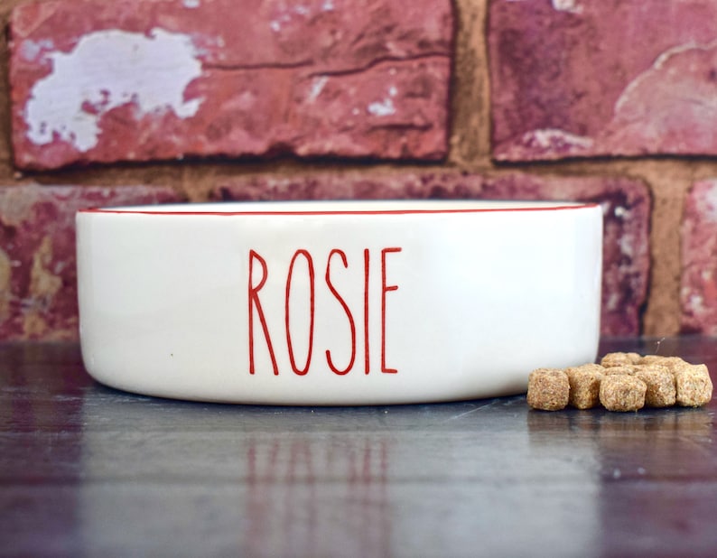 K&ouml;nnte beinhalten: Wei&szlig;e Keramik-Sch&uuml;ssel f&uuml;r Haustiere mit rotem Rand und dem Namen "Rosie" in roten Buchstaben.