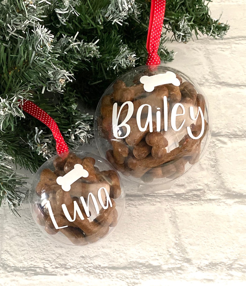 May include: Two clear baubles filled with dog treats. Each bauble has a white bone graphic and a name in white script: "Bailey" and "Luna". Red and white polka dot ribbon hangs from the top of each bauble.