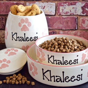 May include: White ceramic pet food bowl and food storage container with pink paw prints and the name Khaleesi written in black. The bowl is filled with brown dog food.