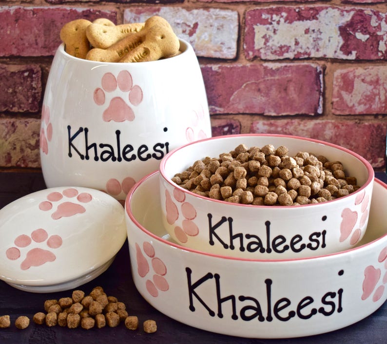 May include: White ceramic pet food bowl and food storage container with pink paw prints and the name Khaleesi written in black. The bowl is filled with brown dog food.