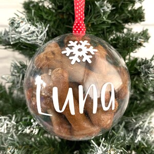 May include: A clear bauble filled with brown treats, suspended by a red and white polka dot ribbon. The bauble features the name "Luna" in white script and a white snowflake. It's displayed on a green Christmas tree, a festive decoration.