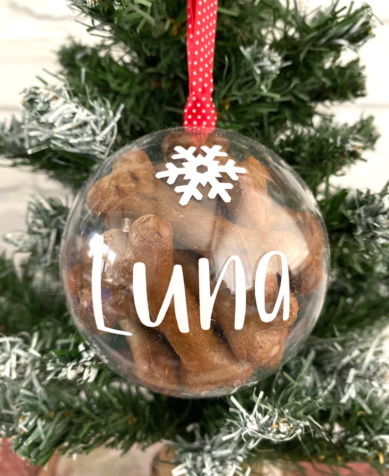 May include: A clear bauble filled with brown treats, suspended by a red and white polka dot ribbon. The bauble features the name "Luna" in white script and a white snowflake. It's displayed on a green Christmas tree, a festive decoration.